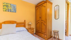 A bedroom with a wooden wardrobe, a bed with a white bedspread, a nightstand with a lamp, and a small stool.