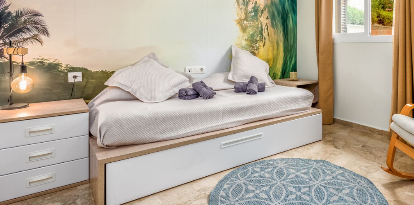 A bedroom with a bed, bedside table, lamp, and a view of greenery through the window.