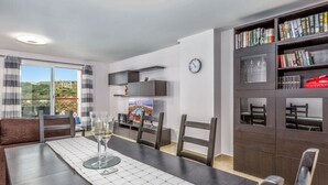 Un coin repas avec une table dressée pour deux personnes, une bibliothèque remplie de livres et d’objets décoratifs, et une vue sur l’extérieur grâce à de grandes fenêtres.