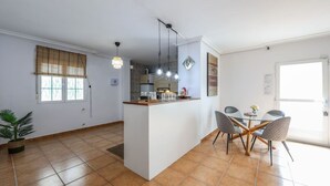 Et moderne køkken med en central ø, hængende lamper og et spiseområde med et glasbord og stole.