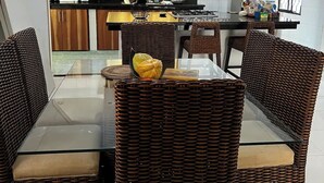 Een eetgedeelte met een glazen tafel en rieten stoelen, een plafondventilator en een keuken op de achtergrond.