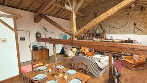 Een landelijke eethoek met een houten tafel gedekt voor een maaltijd, een gezellig woongedeelte met een bank en een boekenkast, en een hoog houten plafond.