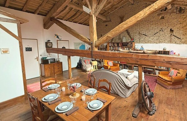 Un coin repas rustique avec une table en bois dressée pour le repas, un espace de vie chaleureux avec un canapé et une bibliothèque, et un plafond en bois particulièrement haut.