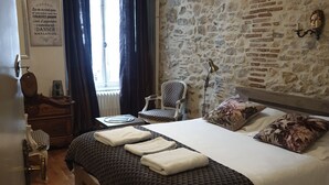 A bedroom with a stone wall, a bed with a patterned bedspread, a chair, a nightstand, and a window with curtains. - L (Saint-Amand-Montrond)