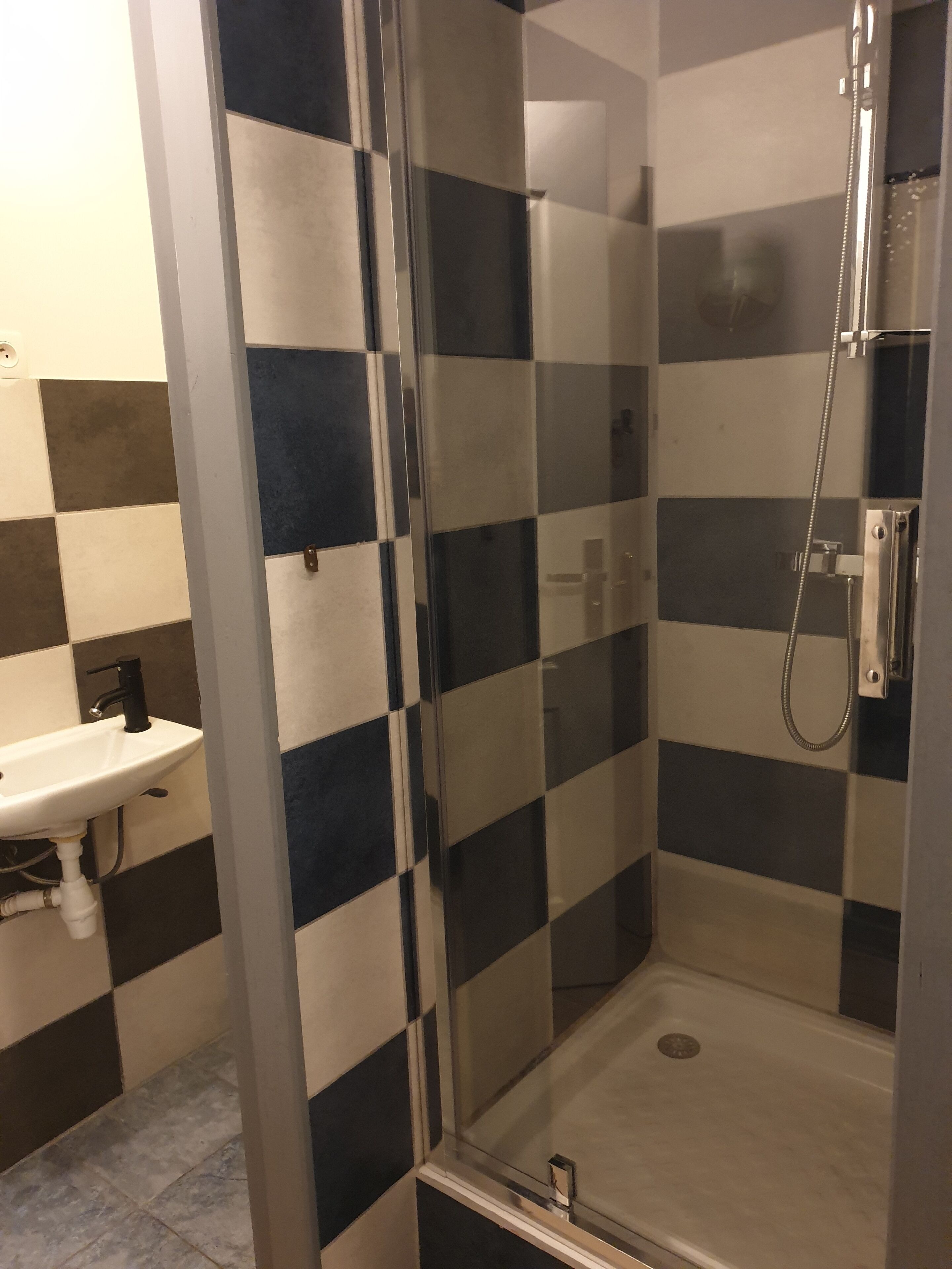 Une douche fermée par des parois de verre, dotée d’un pommeau de douche à main, un lavabo blanc avec un robinet noir et un mur recouvert de carreaux à motifs.