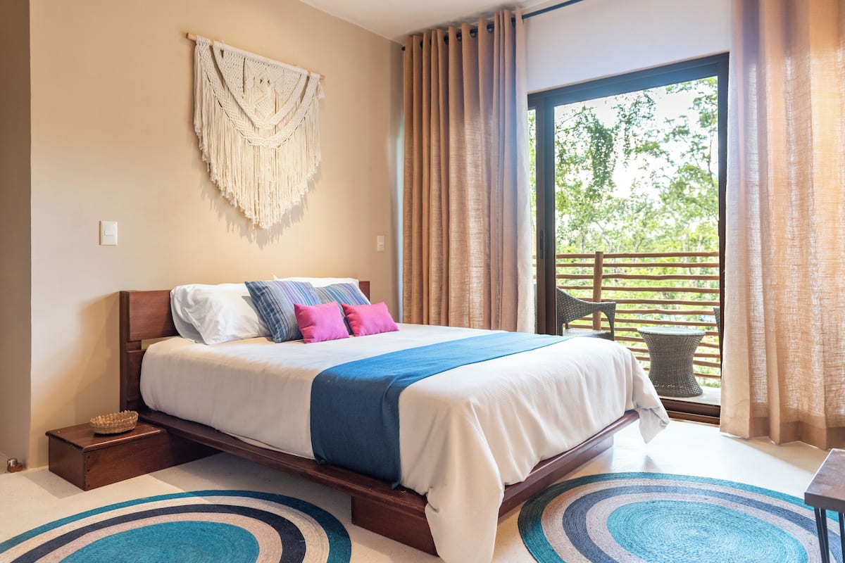 A bedroom with a bed, a macrame wall hanging, and a view of greenery through a window.