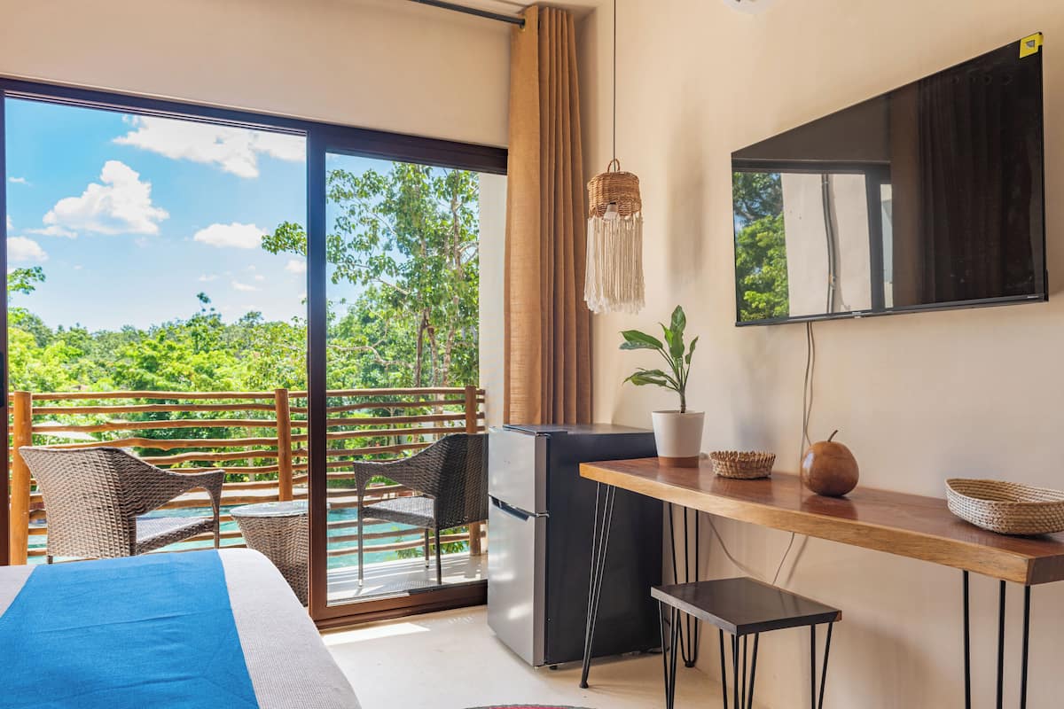 A room with a bed, a desk, a refrigerator, a potted plant, and a view of greenery through a sliding door.