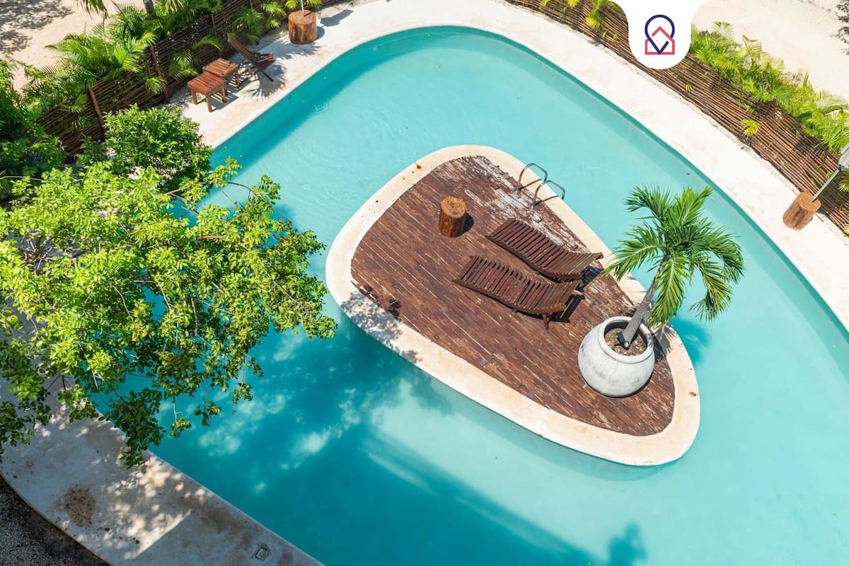 An aerial view of a swimming pool with a wooden deck area, surrounded by lush greenery and a sandy beach.