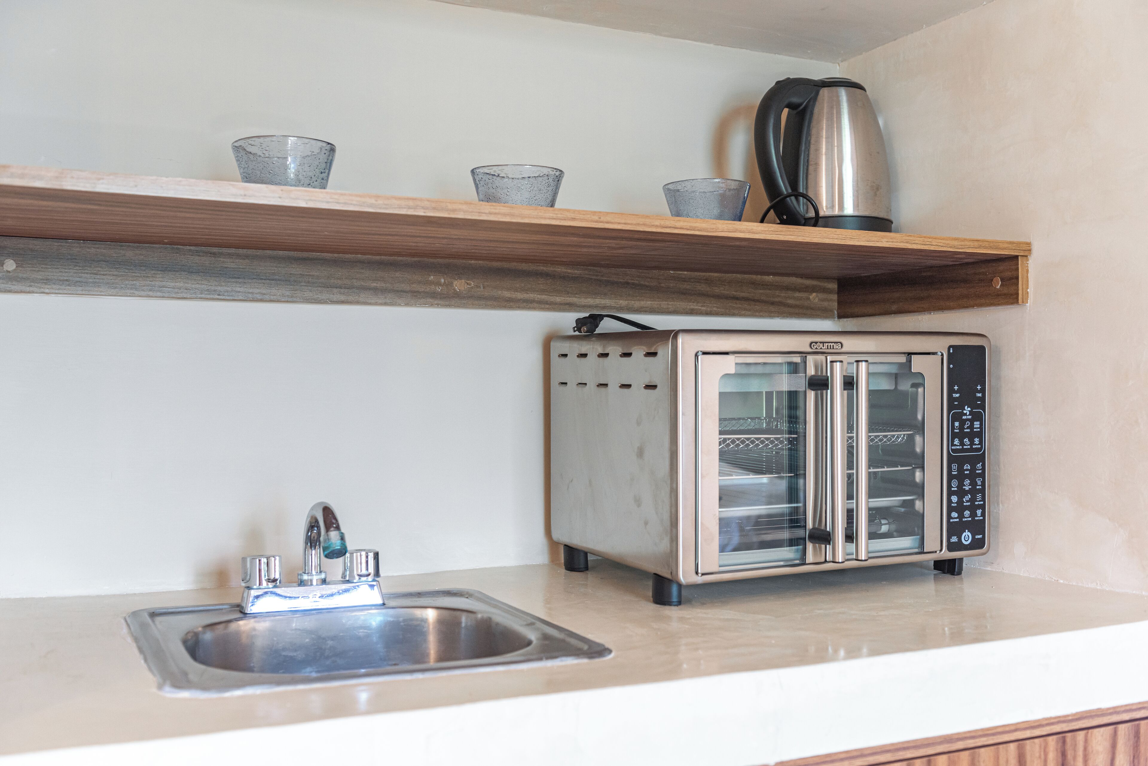 A kitchen with a stainless steel sink, a toaster oven, and a shelf with cups and a kettle.