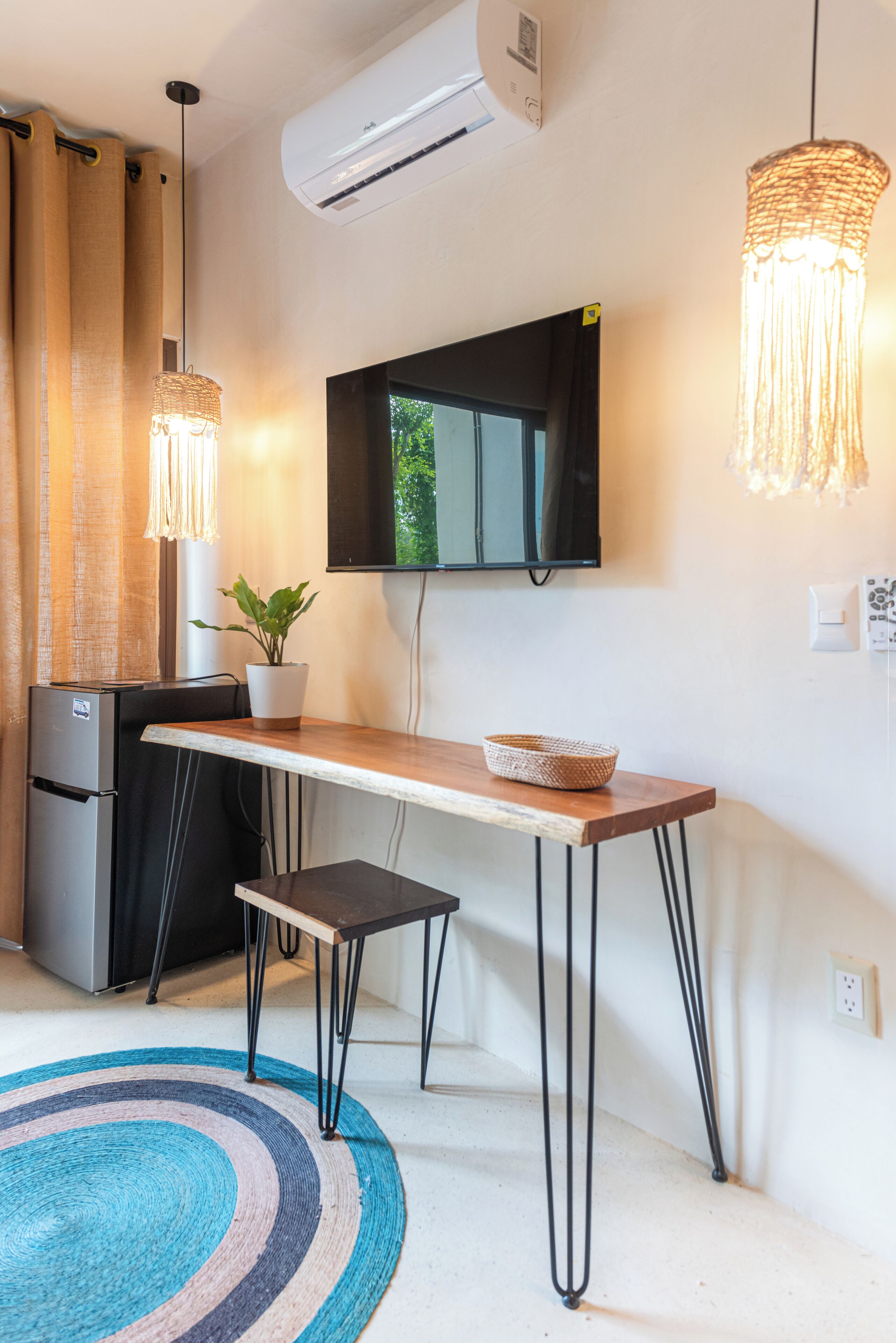 A compact kitchenette with a refrigerator, a small table, and a bench. There is a potted plant on the table, a woven light fixture above, and a flat-screen TV mounted on the wall.