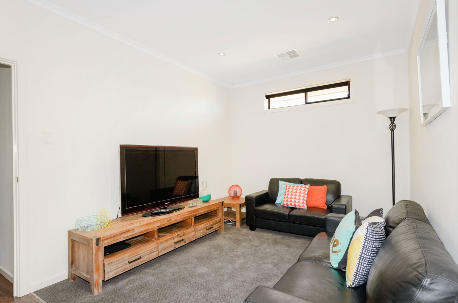 A living room with a flat-screen TV, a wooden entertainment unit, a black leather sofa, and a floor lamp.