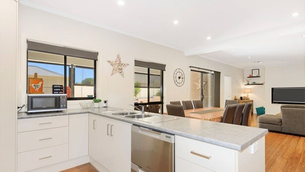 A modern kitchen with white cabinetry, a built-in microwave, and a large island with a sink. There is a dining area with a table and chairs, and a living room with a couch and a flat-screen TV. - Calm Waters, Moonta Bay (Moonta Bay)