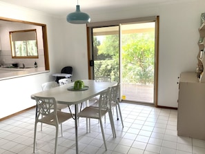 Un comedor con una mesa y sillas blancas, una lámpara colgante azul, una planta de interior pequeña y un divisor corredero de vidrio que da a un patio exterior.