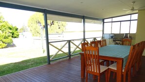 A covered outdoor patio with a wooden table and chairs, a ceiling fan, and a view of a grassy area and trees. - Ardy House, Ardrossan (Ardrossan)