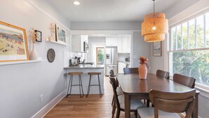 A modern kitchen with white cabinets, a wooden island with stools, and a dining area with a large table and chairs. - Cliff View by Avantstay Hot Tub + Fire Pit 1 Block From Downtown La Jolla (La Jolla)