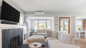 A modern living room with a fireplace, a large flat-screen TV, a sectional sofa, and a dining area visible through an open door. - Cliff View by Avantstay Hot Tub + Fire Pit 1 Block From Downtown La Jolla (La Jolla)