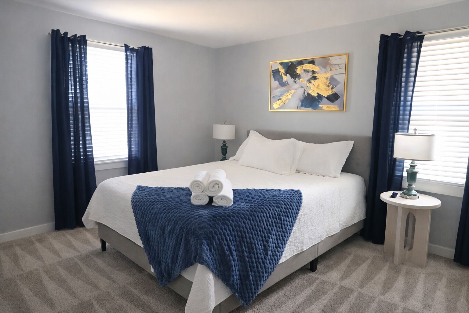 Un letto rifatto con lenzuola bianche e una coperta blu, una lampada da comodino e un quadro appeso al muro.