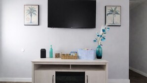 Una sala de estar con un televisor de pantalla plana montado en la pared, un mueble de entretenimiento de color claro con espacio de almacenamiento, un florero con flores azules y dos cuadros enmarcados.