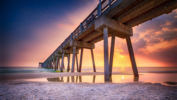 Un muelle se extiende sobre una playa con un atardecer al fondo.