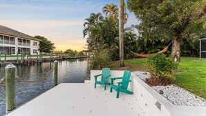 Un patio frente al agua con dos sillas Adirondack verdes, una hamaca y vistas a un puerto deportivo con un edificio al fondo.
