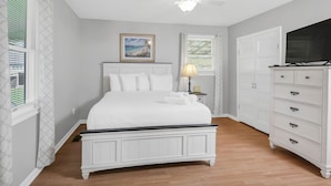Une chambre à coucher comprenant un lit, une table de chevet, une commode, un ventilateur de plafond et une fenêtre avec des stores.