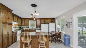 Une cuisine dotée de meubles en bois, d’un îlot central avec deux chaises, d’une table à manger et offrant une vue sur l’extérieur grâce à de grandes fenêtres.