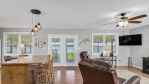 Un vaste espace de vie doté d’un plancher en bois, d’un canapé en cuir, d’une table à manger avec des chaises, d’un éclairage suspendu, d’un ventilateur de plafond et d’un téléviseur fixé au mur.