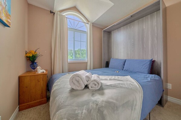 A bedroom with a bed, a nightstand, a window with curtains, and a small vase with flowers.