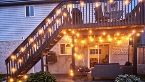 A house with a wooden staircase, a covered porch with string lights, a patio with outdoor furniture, and a well-maintained lawn. - Family friendly summer getaway, short walk to beach, restaurants, etc... (Meaford)