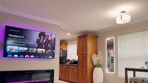 A living room with a flat-screen TV mounted on the wall, a fireplace, wooden cabinets, a dining area with a table and chairs, and a large vase with white flowers. - Beautiful Oasis in Fort Lauderdale (Davie)