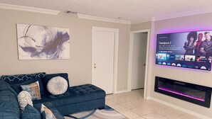 A living room with a dark blue sectional sofa, a large abstract painting, and a flat-screen TV displaying 'Deadpool 2'. - Beautiful Oasis in Fort Lauderdale (Davie)