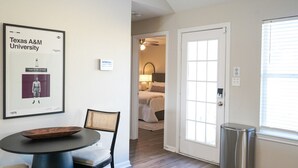 Une chambre d’hôtel avec une table à manger, une chaise, un tableau encadré au mur, une porte donnant sur une autre pièce et une poubelle.