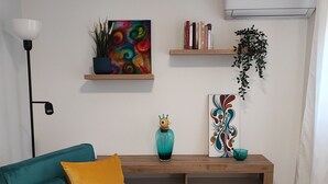 A living room with teal and mustard-colored furniture, a coffee table with a remote and a bowl of chocolates, a colorful abstract painting, a bookshelf, and an air conditioning unit. - Vacation home in the heart of Petralia Soprana with Breakfast (Petralia Soprana)
