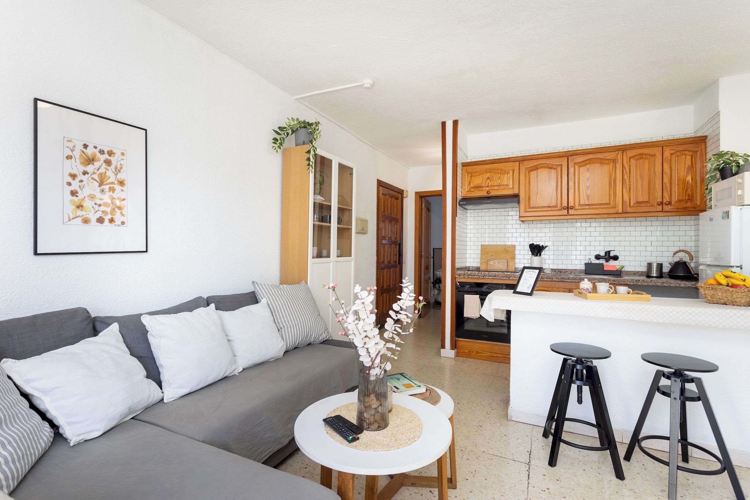 A modern living room with a grey sofa, a small white table with a vase, and a kitchen area with wooden cabinets and a white countertop.