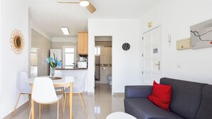 Una sala moderna con un sofá gris, una mesa de comedor con sillas blancas, un ventilador de techo y una cocina al fondo.