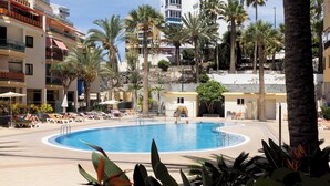 A swimming pool surrounded by palm trees and sun loungers, with a view of multi-story buildings in the background. - Home2Book Cozy Coastal Apt & Pool, Los Cristianos (Costa del Silencio)