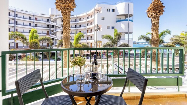 A balcony with a round table, two chairs, and a bottle of wine. There are palm trees and a building with multiple floors in the background. - Home2Book Cozy Coastal Apt & Pool, Los Cristianos (Costa del Silencio)