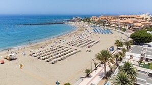 A beach with rows of sun loungers and umbrellas, a clear blue sea, and buildings in the background. - Home2Book Cozy Coastal Apt & Pool, Los Cristianos (Costa del Silencio)