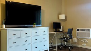 A room with a ceiling fan, a flat-screen TV on a white dresser, a desk with a computer, a chair, and an air conditioning unit. - Charming studio in Newport Beach, fitness gym, pools and more! (Newport Beach)