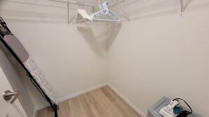 Een kleine, verzorgde kamer met een houten vloer, een witte deur, een metalen kapstok en een klein opbergkastje met een stekkerdoos.
