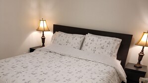 Een net opgemaakt bed met een bloemenprint deken en kussens, aan weerszijden geflankeerd door nachtkastjes met lampen, tegen een effen muur en houten vloer.