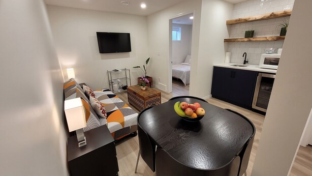 Een moderne woonkamer met een bank, salontafel en een aan de muur bevestigde televisie. Er is een klein keukenhoekje met een magnetron en een spoelbak.