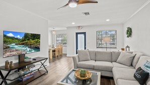 A modern living room with a large flat-screen TV, a ceiling fan, a sofa, a coffee table with a plant, and a dining area in the background.