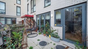 A residential building with a brick exterior, large windows, and a garden area with potted plants and outdoor furniture.