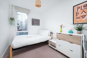 A bedroom with a bed, bedside table, dresser, window, hanging plant, and artwork.