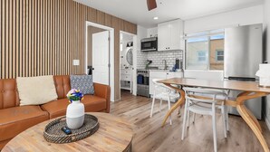 A modern living room with a brown leather sofa, a wooden dining table with white chairs, and a ceiling fan.