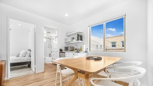 A modern, well-lit apartment with a wooden dining table and white chairs, a kitchen with white cabinets, and a large window offering a view of buildings.