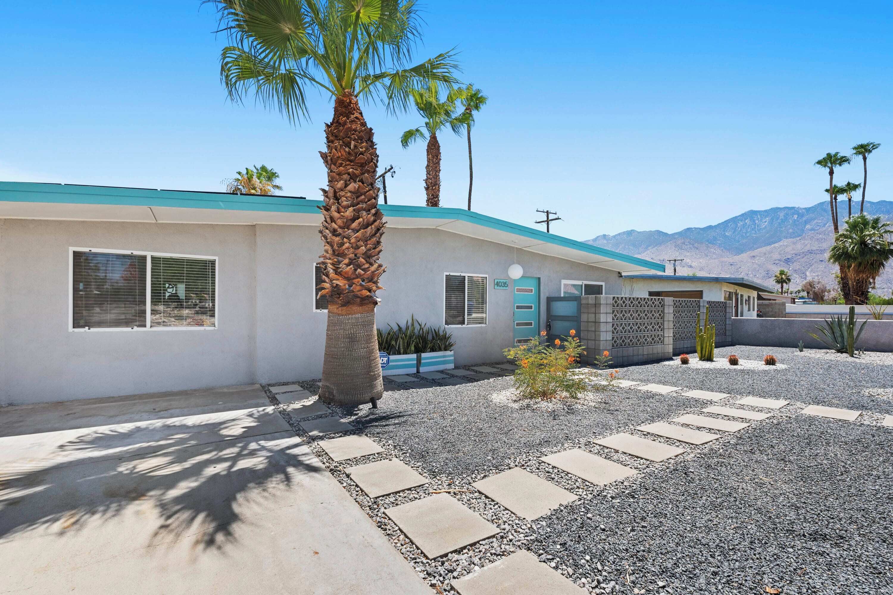 A modern two-story house with a light-colored exterior, a blue roof, and a gravel driveway with stepping stones.