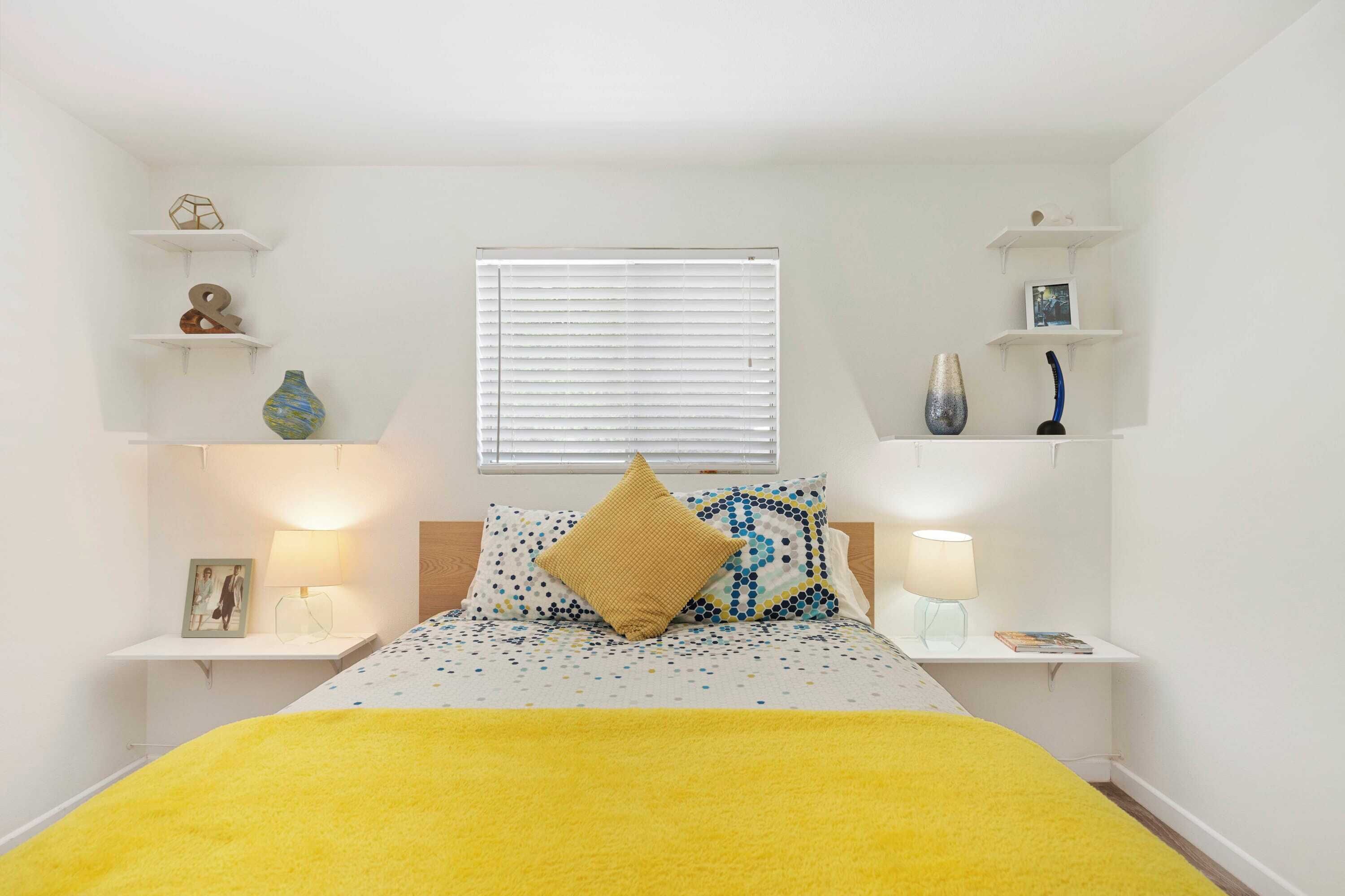 A bedroom with a bed, bedside tables, a window with blinds, and decorative items on shelves.