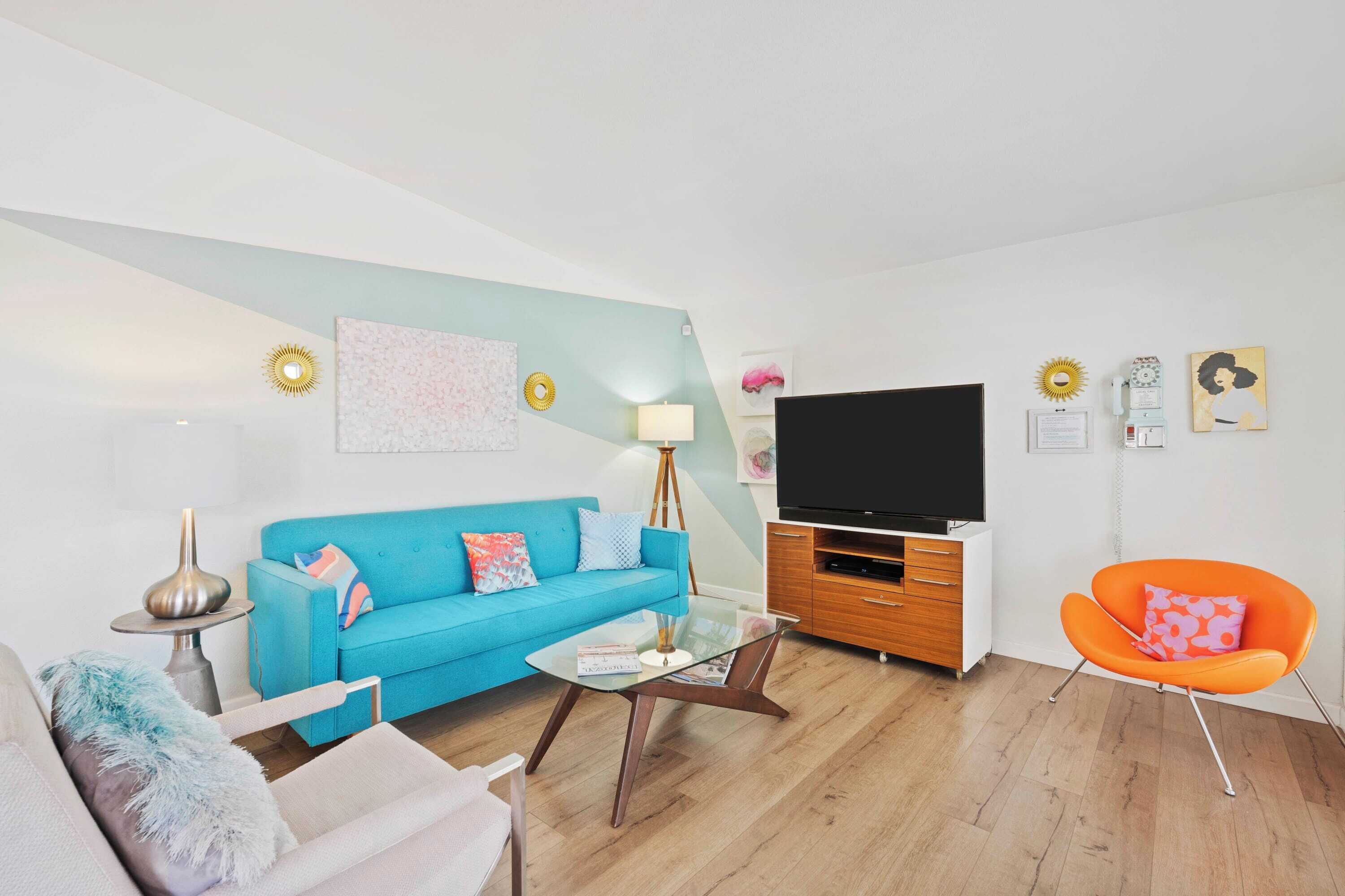 A modern living room with a blue sofa, a glass coffee table, a wooden TV stand, and an orange armchair.